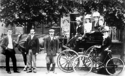 Cole family, outside Cole's Dairy, setting off on an outing?
Three ladies in one-horse carriage, one lady with bicycle. One man driving carriage, three men standing at side of horse. Smartly dressed with buttonholes, moustaches, hats, pipes. Shop or restaurant in background has advertisements for R. White's Crown cyder, ginger ale, Cadbury's chocolate, R. White's lemon squash and American cream soda.
Keywords: bicycles;horse-drawn vehicles;horses;shops;caterers;advertisements;transport;road transport
