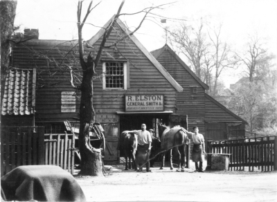 Smithy at corner of St. George's Road, Forty Hill
Weatherboarded building with signboard reading "R. ELSTON / GENERAL SMITH &c. / HORSES CAREFULLY SHOD". Two men with aprons stand outside, each with one hand on a horse and the other holding a hammer.
Keywords: 1910s;weatherboarding;blacksmiths;farriers;horses