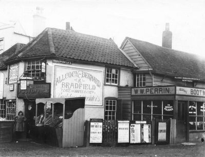 Post Office, Palmers Green
Post Office on the corner of Hazelwood Lane. The Post Office has a large advertisement on the side wall, reading "Allcock-Bernard & Bradfield / (Late of Maple & Co. Ltd) / Builders & Decorators". Their weatherboarded workshop is adjacent, behind the shop of "W. W. Perrin, Bespoke boot[maker]". The lamp hanging on the corner outside the Post Office door has the legend "YE OLDE PALMERS GREEN REGISTRY OFFICE ...". A poster on the fence, from Bowes Park Congregational Church, announces that the Rev. Henry Varley, B.A., will preach on Sunday next on "The gospel of the empty tomb". 
Keywords: post offices;shops;weatherboarding