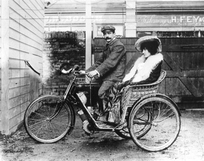 Motorcycle and sidecar
Large twisted bulb horn on handlebars; motor inscribed "MINERVA ..." on crankcase. Location unknown, though shop behind has sign "Fishmongers H. FEY & Co."

William John Graham and his brother Sydney were in business making bicycles until the motor car age arrived when they started as garage proprietors in premises at 56 Church Street, Enfield.  This was the shop and offices; the workshops were at the rear in Sarnesfield Rd.  WJ Graham actually registered the first patent in 1903 for the motorcycle sidecar.

The picture shows Sydney Graham and his wife Beatrice in a Graham Bros Sidecar circa 1906. 

The other business in the background appears to be "JW Booth"

Keywords: bicycles;road transport;motorcycles